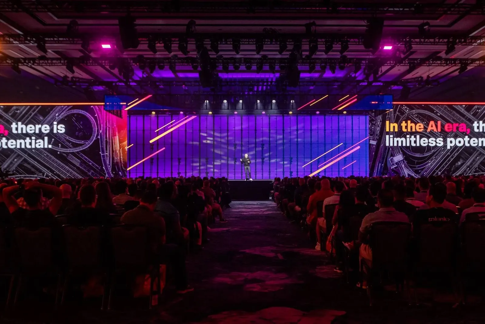 Keynote speaker on a conference stage in front of a massive purple LED wall with a full audience, photographed by Marquee Visuals.