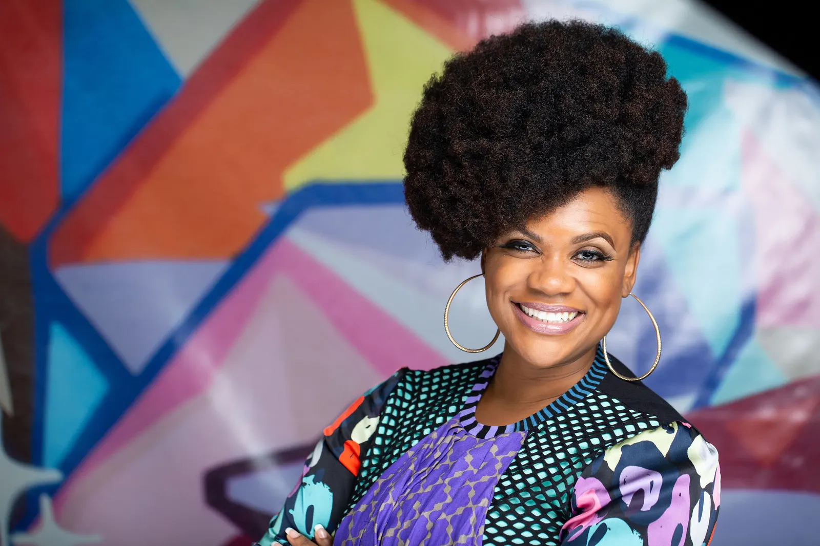 Corporate headshot of a smiling woman with a large curly updo and a colorful floral top against a vivid mural backdrop, by Marquee Visuals in Los Angeles.