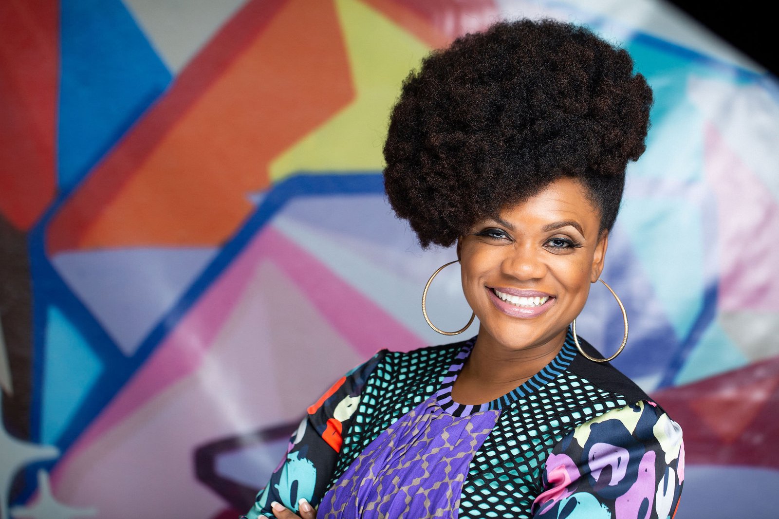 Corporate headshot of a smiling woman with a large curly updo and a colorful floral top against a vivid mural backdrop, by Marquee Visuals in Los Angeles.