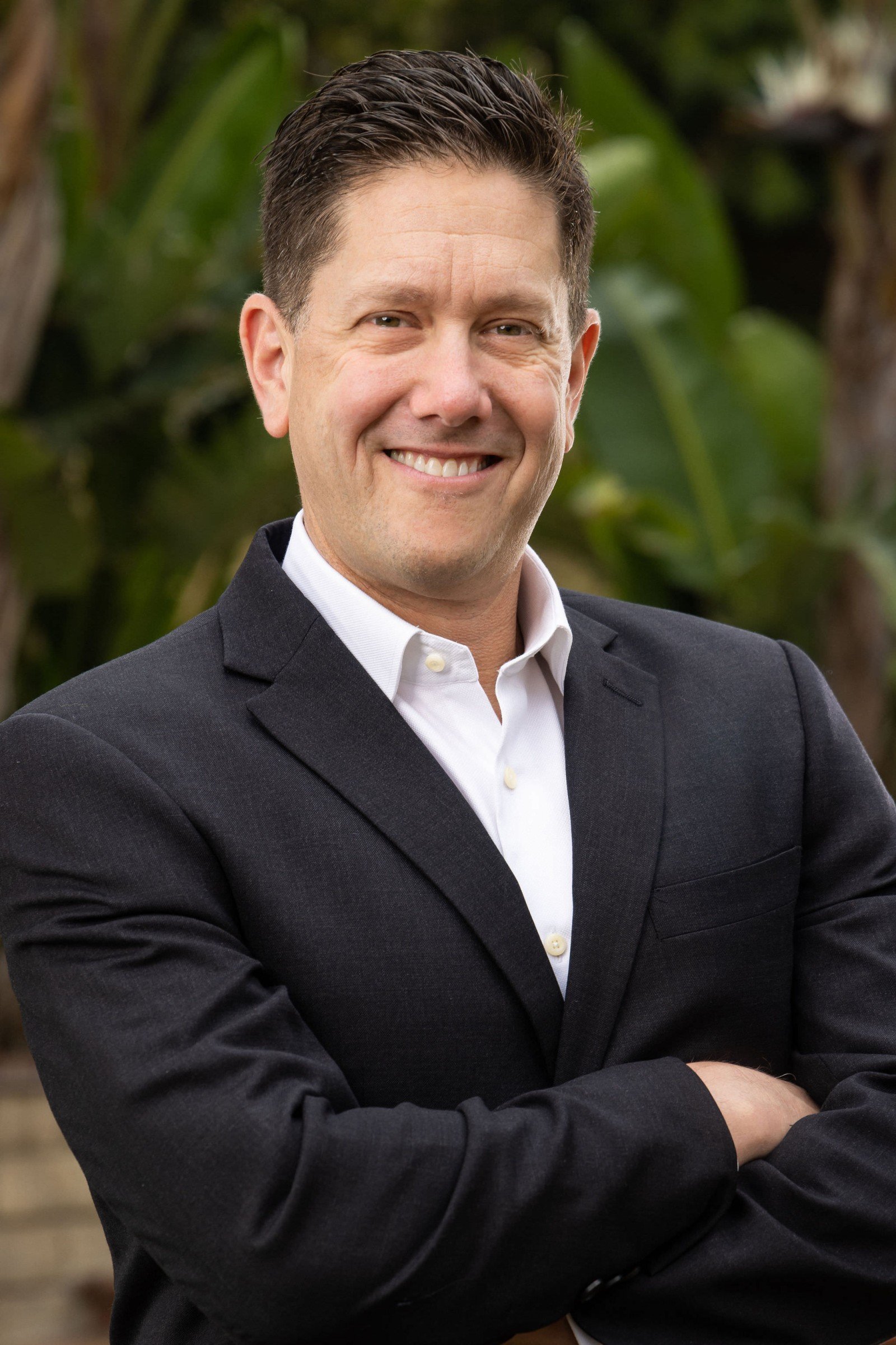 Executive headshot of a man with glasses in a dark suit, arms crossed, photographed outdoors with a soft green bokeh backdrop, by Marquee Visuals in Los Angeles.
